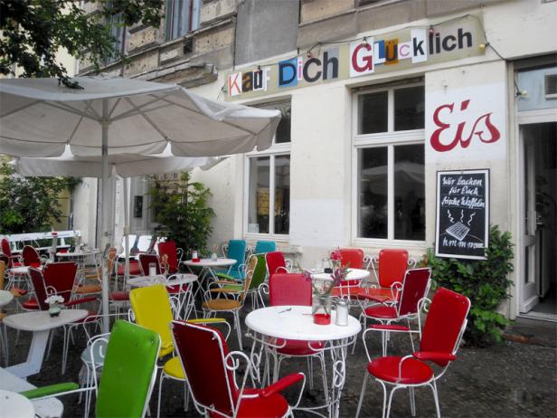 Einladende Außenterrasse eines Cafés mit bunten Stühlen und Tischen unter einem Sonnenschirm.