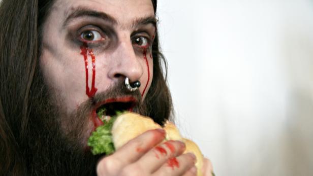Ein Mann mit blutverschmiertem Gesicht und Nasenring isst ein Sandwich.