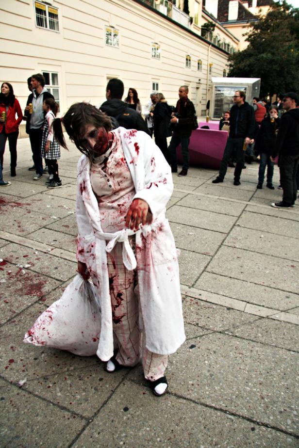 Eine als Zombie verkleidete Frau mit blutverschmiertem Bademantel steht auf einem Platz.