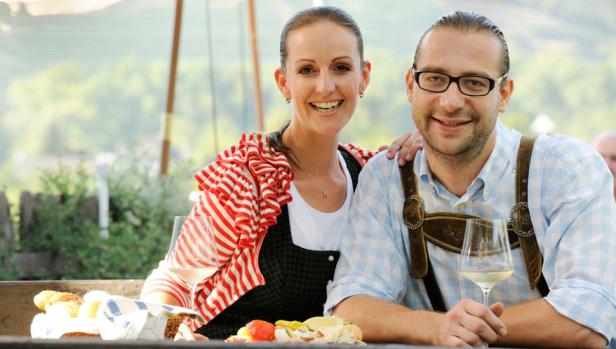 Ein Paar in Tracht sitzt an einem Tisch mit Brot, Wurst und Wein.