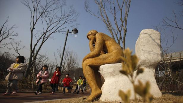 Kinder spielen im Park neben einer Skulptur, die an „Der Denker“ erinnert.
