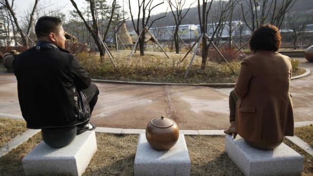 Ein Mann und eine Frau sitzen auf Steinsockeln in einem Park, zwischen ihnen steht ein Gefäß.