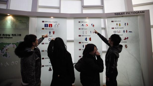 Eine Gruppe von Menschen betrachtet eine Ausstellung von Toiletten-Piktogrammen verschiedener Länder.