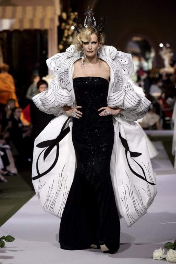 Eine Frau in einem schwarzen, trägerlosen Kleid und einem weißen Mantel mit übergroßen Ärmeln auf einem Laufsteg.