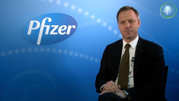 Ein Mann sitzt vor einem blauen Hintergrund mit dem Pfizer-Logo.