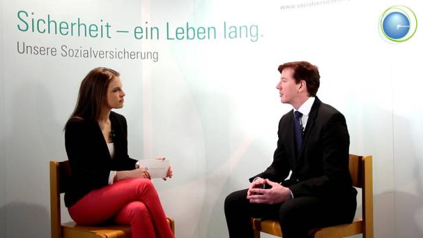 Eine Frau interviewt einen Mann vor einem Hintergrund mit dem Slogan „Sicherheit – ein Leben lang“.