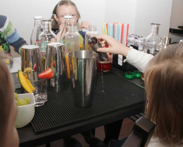 Ein Kind gießt Flüssigkeit in einen Cocktailshaker, umgeben von Früchten und anderen Kindern.