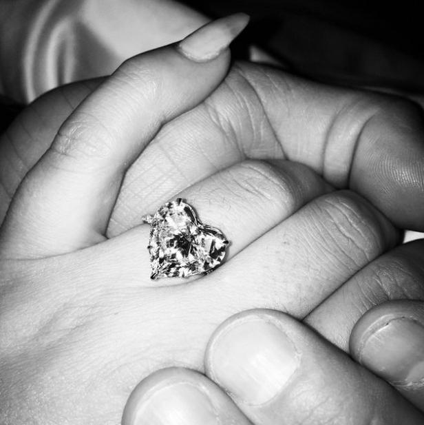 Ein herzförmiger Diamantring an einem Finger, gehalten von einer anderen Hand.