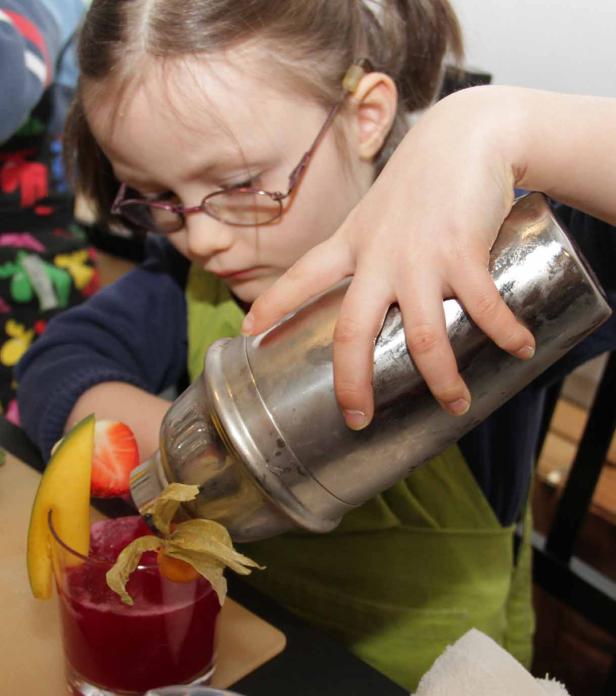 Ein Mädchen mit Brille mixt einen Cocktail mit Früchten in einem Shaker.