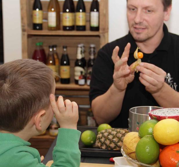 Ein Mann erklärt einem Kind eine Physalis vor einem Hintergrund mit Getränkeflaschen.
