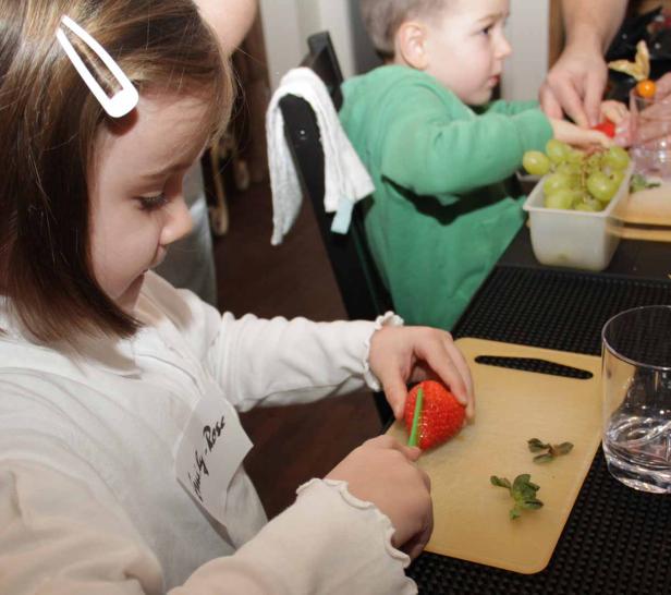 Ein Mädchen schneidet eine Erdbeere, während ein Junge im Hintergrund ebenfalls Obst zubereitet.