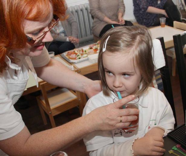 Ein kleines Mädchen trinkt mit einem Strohhalm aus einem Glas, während eine Frau ihr hilft.