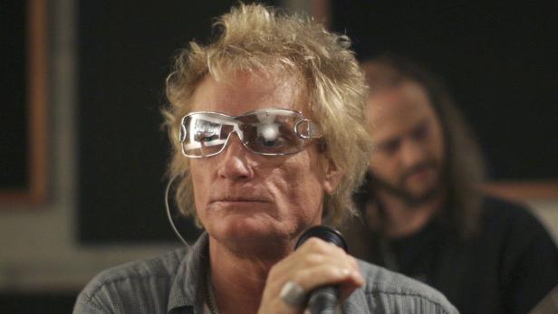 Ein Mann mit blonden Haaren und futuristischer Sonnenbrille hält ein Mikrofon.