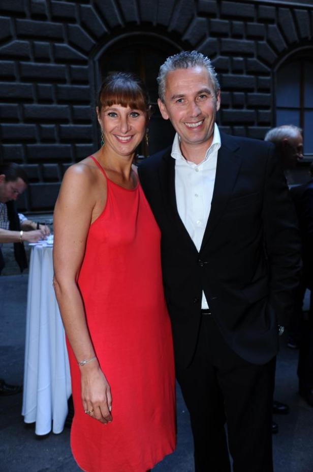 Ein Mann im Anzug und eine Frau in einem roten Kleid posieren lächelnd nebeneinander.