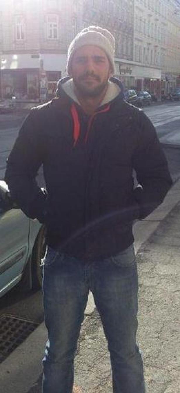 Ein Mann mit Mütze und Jacke steht auf dem Bürgersteig einer Stadtstraße.