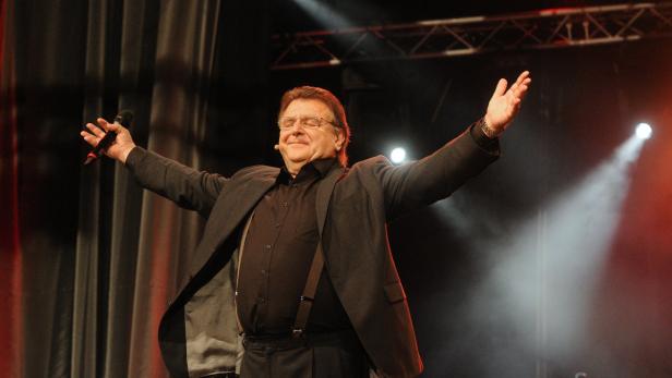 Ein Mann mit Brille steht mit erhobenen Armen auf einer Bühne.