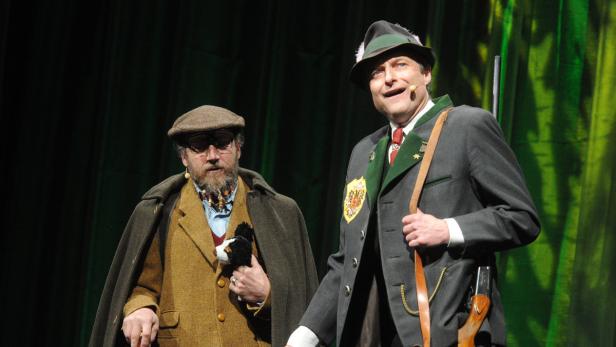 Zwei Männer in Trachten stehen auf einer Bühne vor einem grünen Vorhang.