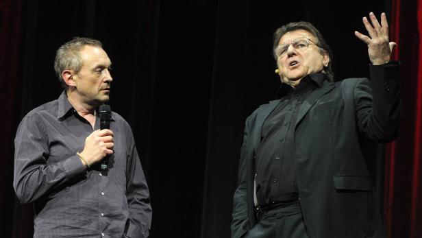 Zwei Männer stehen auf einer Bühne, einer spricht ins Mikrofon, der andere gestikuliert.