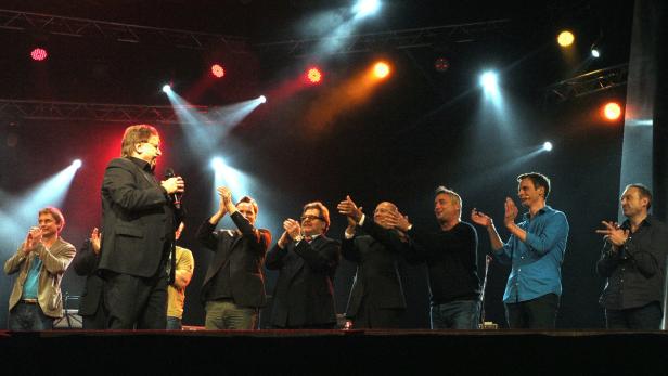 Eine Gruppe von Männern steht auf einer Bühne und applaudiert einem Redner mit Mikrofon.