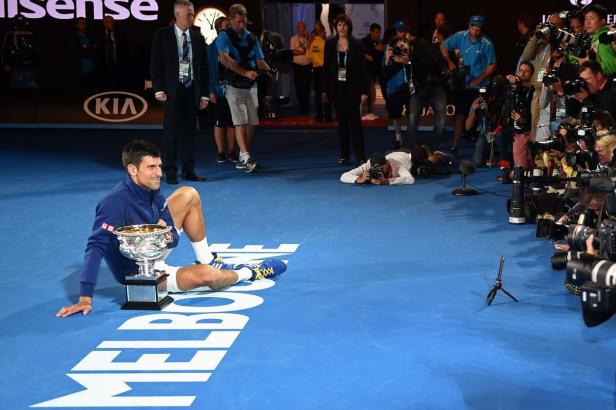 Novak Đoković sitzt mit der Trophäe auf dem blauen Tennisplatz, umringt von Fotografen.