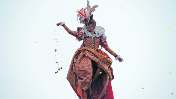 Eine Frau in historischer Kleidung schwebt an einem Seil hängend in der Luft und wirft Konfetti.