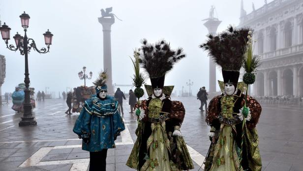 Drei Personen in aufwendigen Kostümen und Masken auf dem Markusplatz in Venedig im Nebel.