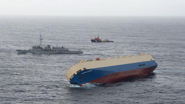 Das Frachtschiff „Modern Express“ liegt Schlagseite im Meer, während ein Schlepper und ein Kriegsschiff in der Nähe sind.