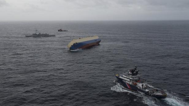 Ein havariertes Frachtschiff liegt auf der Seite im Meer, umgeben von Rettungsschiffen.
