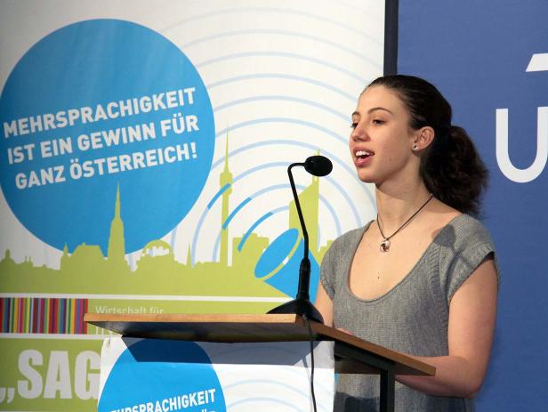 Eine junge Frau hält eine Rede vor einem Hintergrund mit der Aufschrift „Mehrsprachigkeit ist ein Gewinn für ganz Österreich!“.