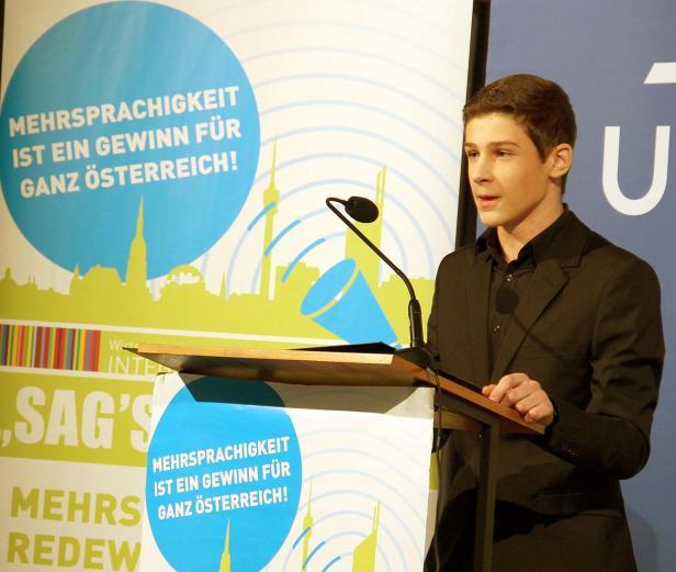 Ein junger Mann hält eine Rede vor einem Hintergrund mit der Aufschrift „Mehrsprachigkeit ist ein Gewinn für ganz Österreich!“.