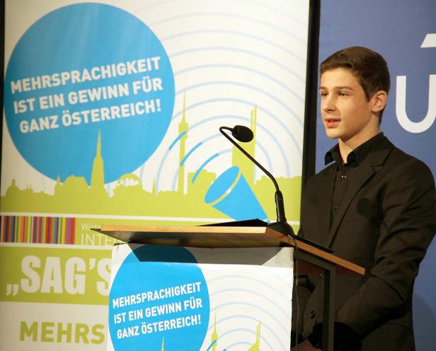 Ein junger Mann steht an einem Rednerpult vor einem Plakat, das Mehrsprachigkeit bewirbt.
