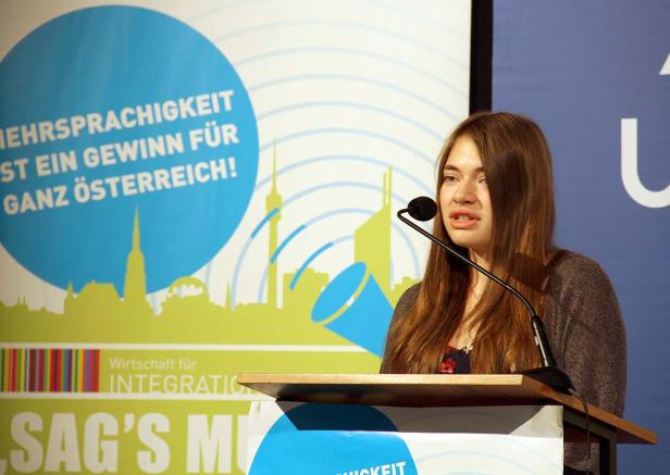 Eine junge Frau spricht an einem Rednerpult vor einem Banner zum Thema Mehrsprachigkeit in Österreich.