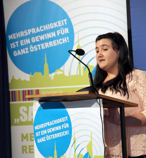 Eine Frau spricht an einem Rednerpult mit dem Slogan „Mehrsprachigkeit ist ein Gewinn für ganz Österreich!".