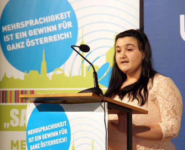 Eine Frau spricht an einem Rednerpult vor einem Hintergrund mit der Aufschrift „Mehrsprachigkeit ist ein Gewinn für ganz Österreich!“.