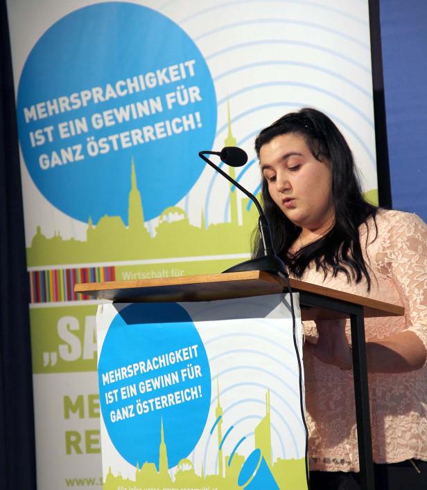 Eine Frau hält eine Rede vor einem Banner, auf dem „Mehrsprachigkeit ist ein Gewinn für ganz Österreich!“ steht.