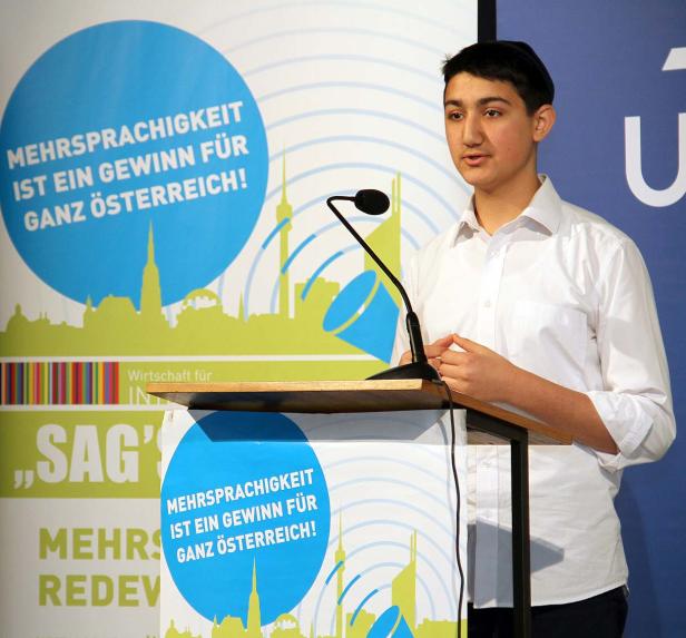 Ein junger Mann hält eine Rede vor einem Banner, das Mehrsprachigkeit in Österreich thematisiert.