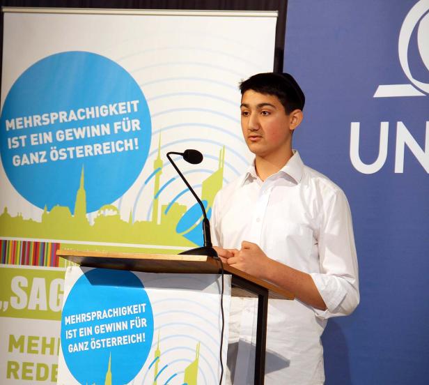 Ein junger Mann steht an einem Rednerpult vor einem Banner mit der Aufschrift „Mehrsprachigkeit ist ein Gewinn für ganz Österreich!“.