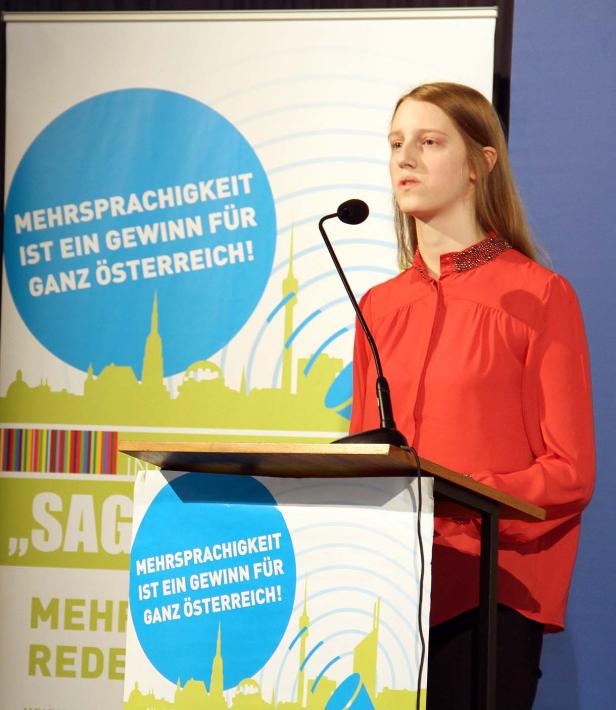 Eine junge Frau hält eine Rede vor einem Banner, das Mehrsprachigkeit bewirbt.