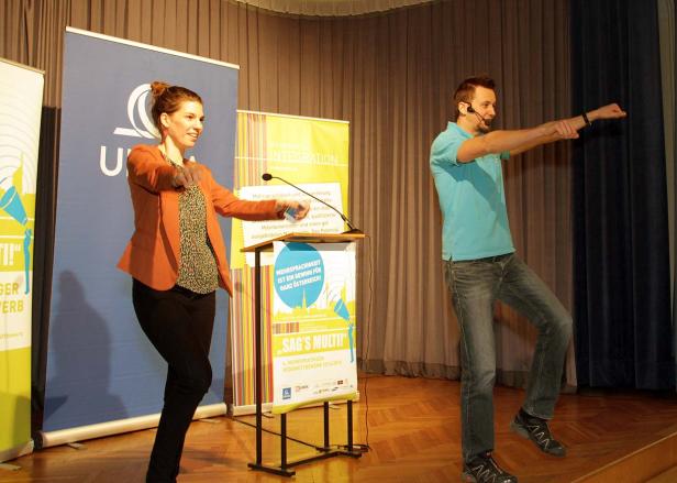 Eine Frau und ein Mann gestikulieren auf einer Bühne während einer Präsentation von „Sag's Multi!“.