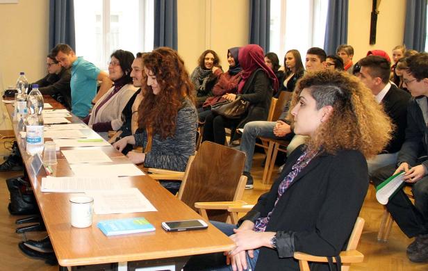 Eine Gruppe von Menschen sitzt in einem Raum an Tischen und hört zu.