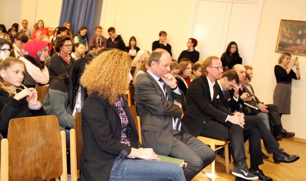 Ein Publikum sitzt auf Stühlen in einem Raum und hört aufmerksam zu.