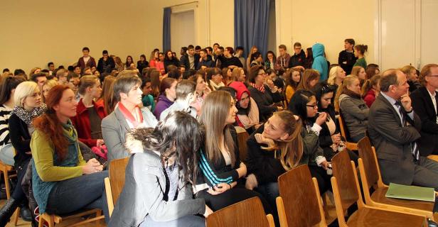 Ein Publikum sitzt auf Holzstühlen in einem Raum und schaut in eine Richtung.