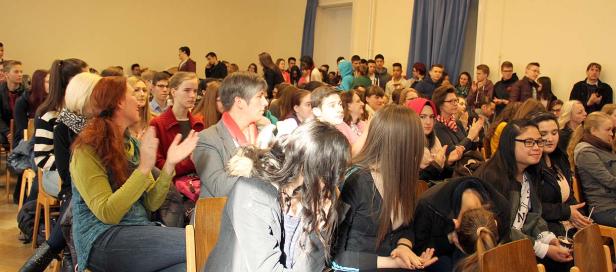 Ein Publikum sitzt auf Stühlen und applaudiert in einem Raum.