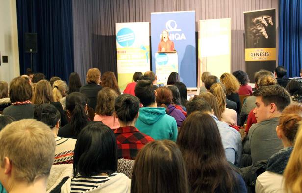 Eine Rednerin steht vor Publikum auf einer Bühne mit Werbebannern.