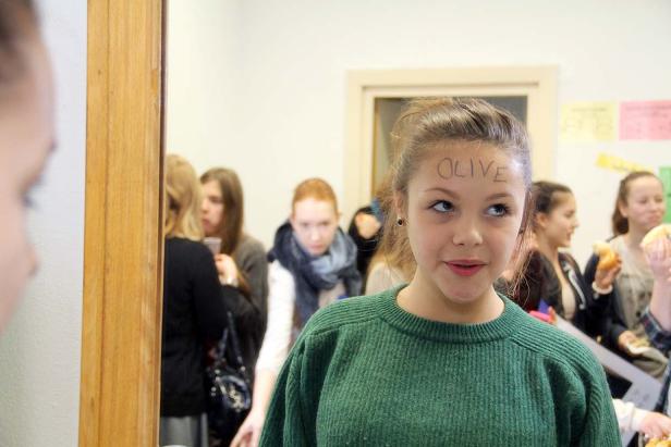 Ein junges Mädchen mit dem Wort „Olive“ auf der Stirn, umgeben von anderen Jugendlichen.