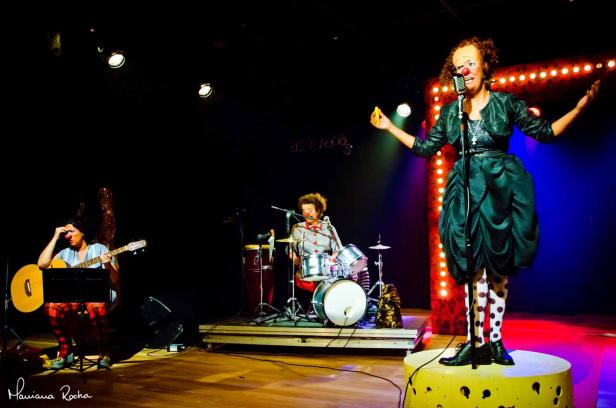 Eine Clownin steht auf einer Bühne und singt mit Band.