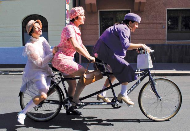 Drei Frauen in Vintage-Kleidung fahren gemeinsam auf einem Tandem-Fahrrad.