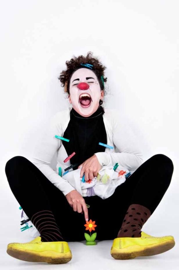 Eine als Clown geschminkte Person mit Wäscheklammern und gelben Schuhen zeigt auf eine Blume.