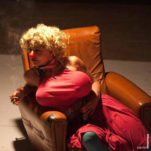 Eine Frau mit Clownsnase und rotem Kleid sitzt in einem Ledersessel und raucht eine Zigarette.