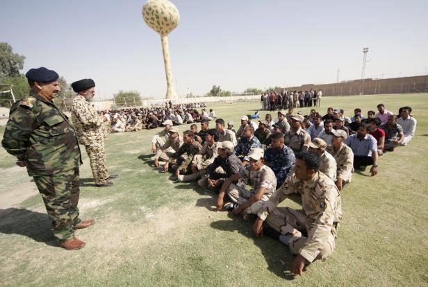 Eine Gruppe von Soldaten sitzt auf einem Rasen und hört zwei Offizieren zu.
